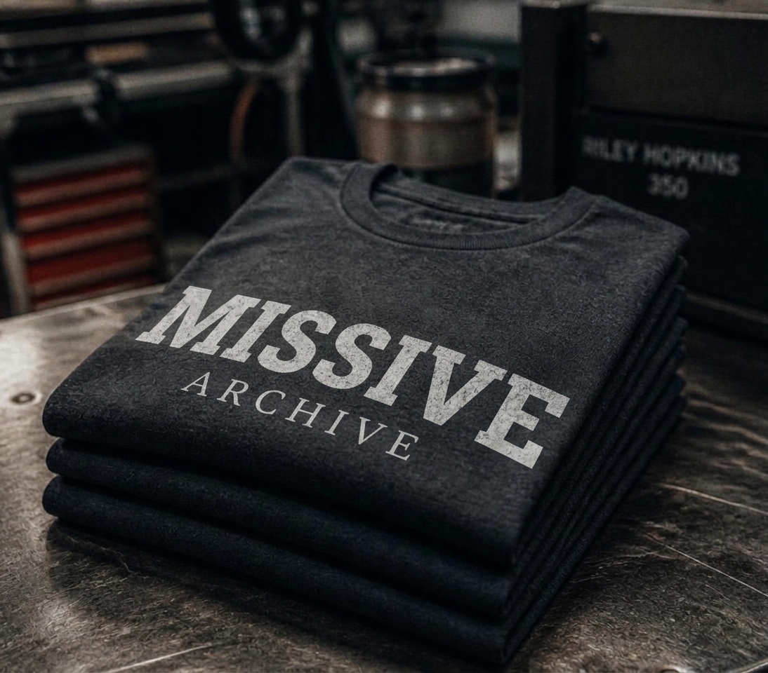Stack of folded black Missive Archive t-shirts with a distressed serif logo print on a steel table